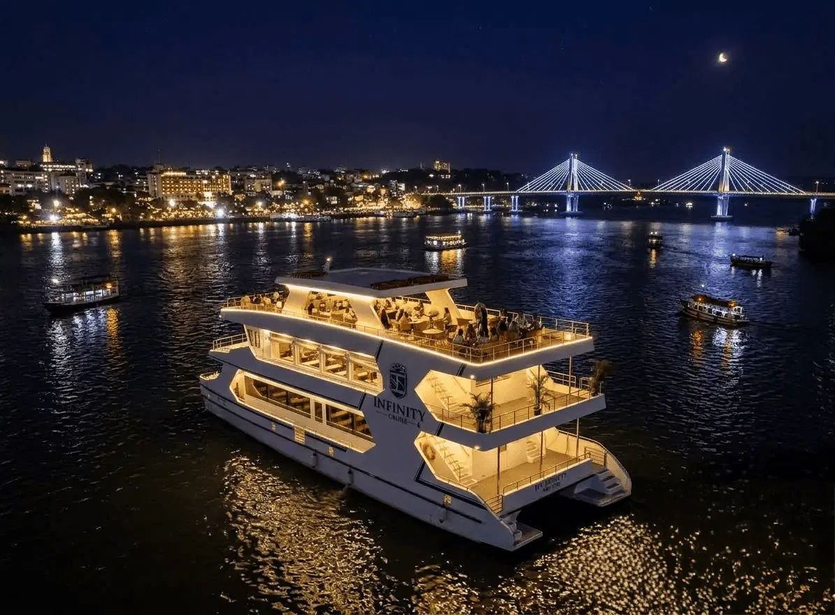 Lit three-deck dinner cruise boat sailing the Mandovi river at night with the bridge and city lights behind it